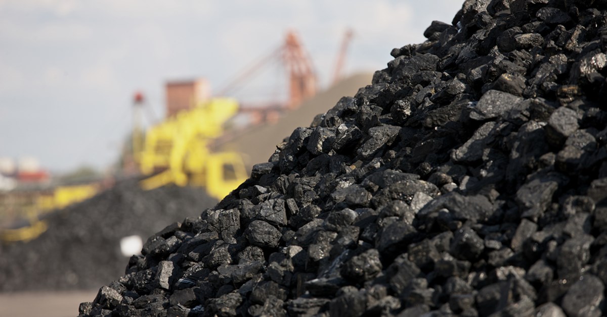 A close-up of a large coal pile with industrial machinery in the blurred background