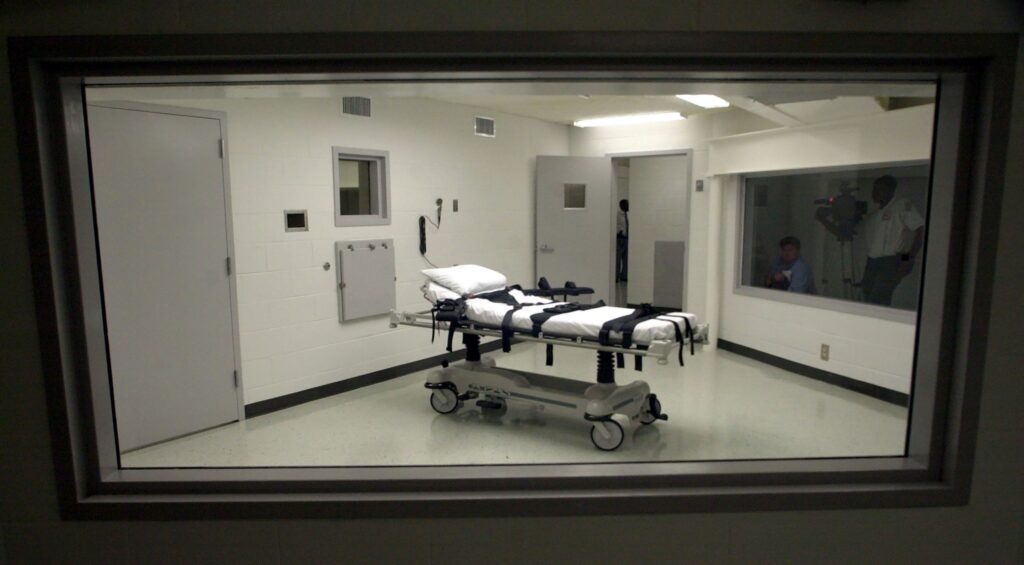 An observation window showing an empty execution chamber with a gurney and restraints, and reflections of observers in the glass.