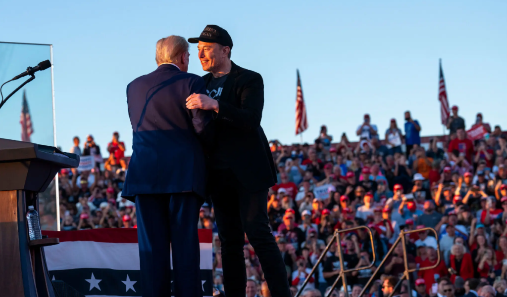 Donald Trump and Elon Musk embracing on stage at a political rally with a crowd of spectators in the background and American flags visible.