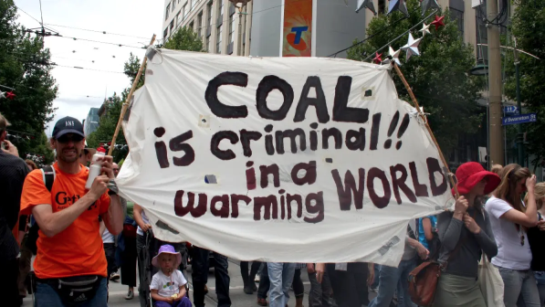 People holding a protest banner that reads "COAL is criminal!! in a WARMING WORLD" on a city street.