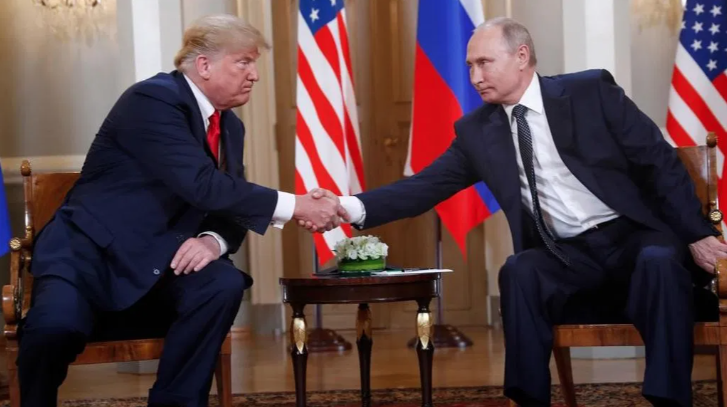 Two political figures seated and shaking hands with flags of the USA and Russia in the background.