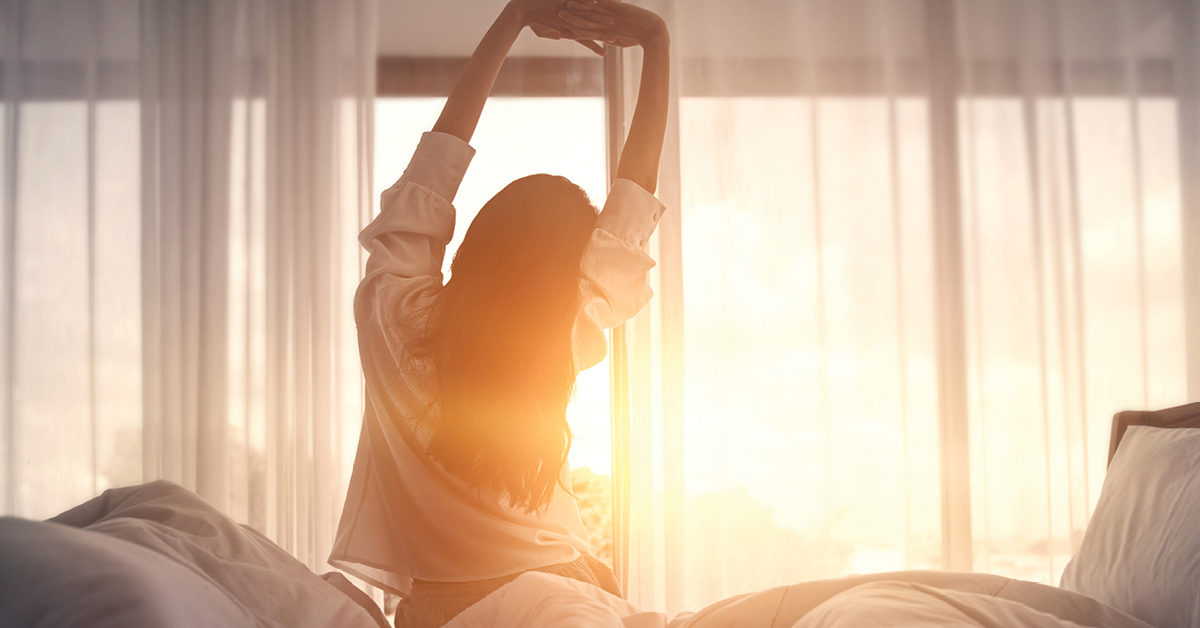 A woman stretching in bed after waking up, sunlight streaming through the curtains