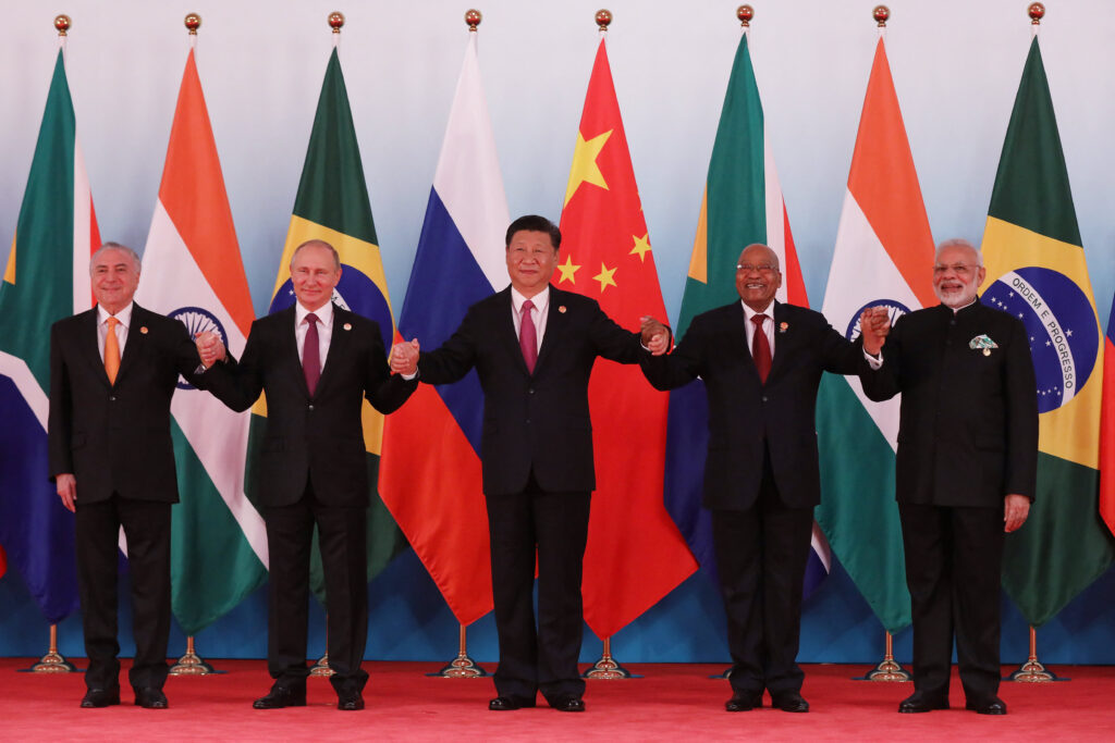 Five world leaders standing in a row, holding hands in front of their countries' flags at an international summit.