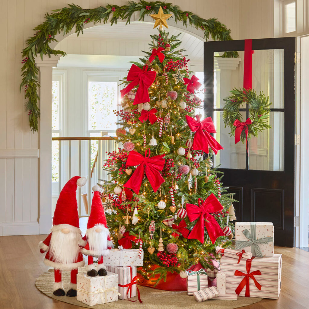 A beautifully decorated Christmas tree with red bows, a golden star, and surrounded by wrapped presents and two gnome figures in a home interior setting.