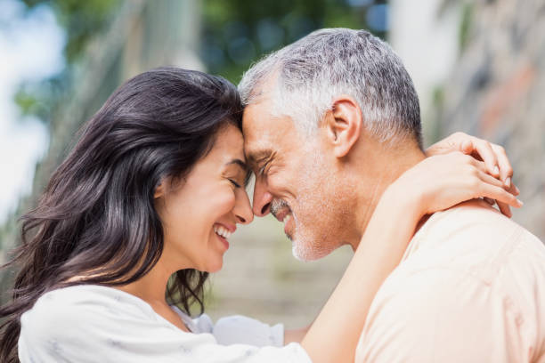 A man and woman share a tender moment, smiling and leaning their foreheads together in a loving embrace, surrounded by greenery and stone steps.