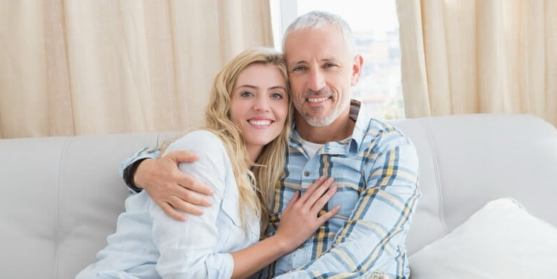 A happy couple sitting closely together on a couch, smiling and enjoying each other's company in a bright, cozy living space.