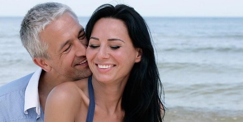 A couple shares a tender moment at the beach, with the man gently embracing the woman, both smiling happily against the backdrop of the ocean.