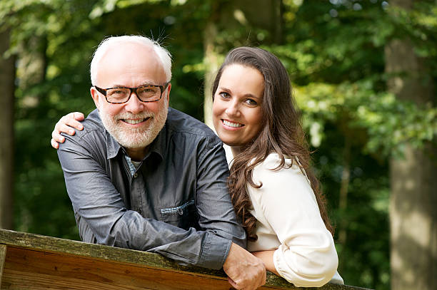 A joyful older man with glasses poses affectionately with a younger woman outdoors, both smiling and embracing each other in a natural setting.