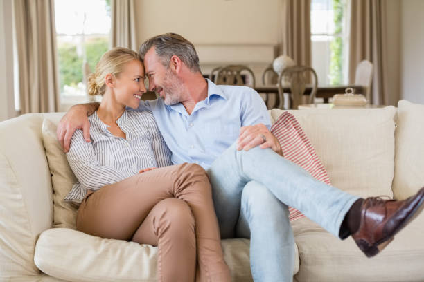 A couple sharing a tender moment while sitting close together on a sofa, smiling and enjoying each other's company in a cozy living room setting.