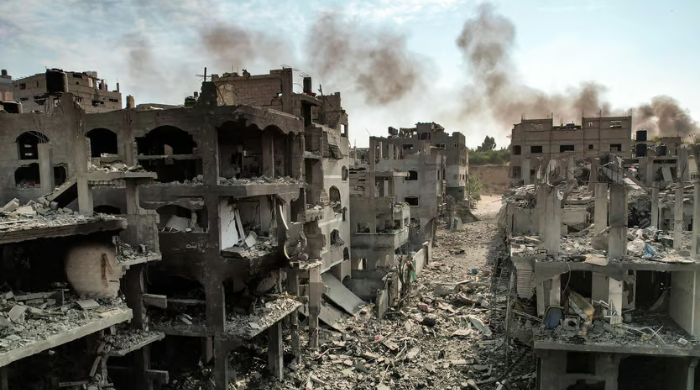 A devastated urban area with heavily damaged buildings and debris, smoke rising in the background. Israel and Gaza