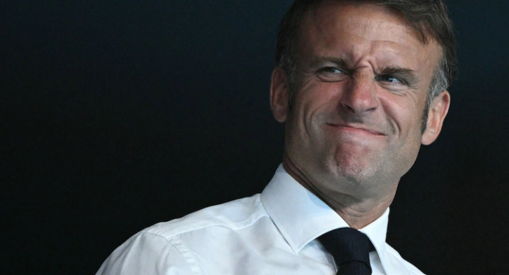 A man in a white shirt and dark tie smirking against a dark background.