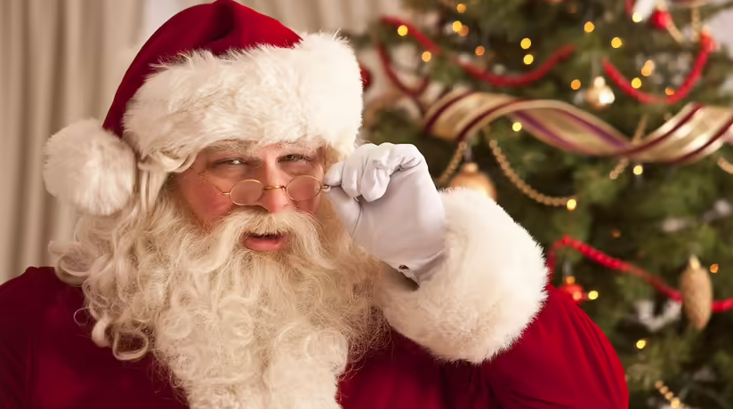 A person dressed as Santa Claus holding his glasses with a Christmas tree in the background.