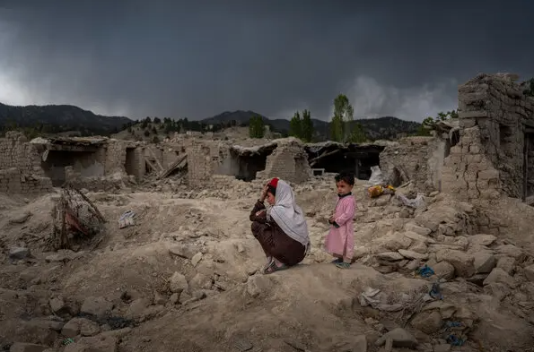 A woman and child stand amidst the ruins of destroyed buildings under a dark sky, conveying a sense of loss and hope.