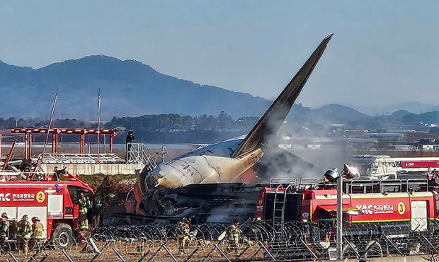 Emergency services tackle a plane crash with smoke rising from the wreckage, showcasing fire trucks and responders at the scene.