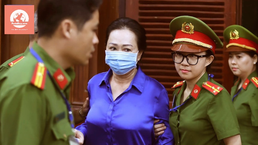 A person in a blue shirt wearing a mask being escorted by uniformed officers.