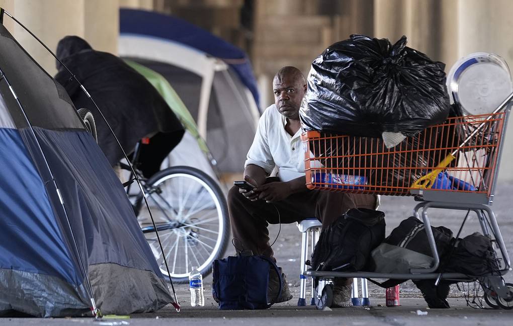 A man sitting on a stool beside a shopping cart filled with belongings, surrounded by tents in a sheltered area.