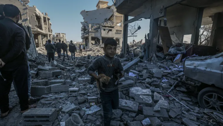 A young boy carrying two kittens amidst the rubble of destroyed buildings, with people in the background surveying the damage.