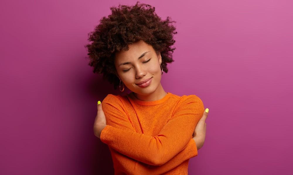 Fifty Ways to Practice Self-Care: How to Pamper Yourself for Any Occasion 2 A young woman smiling softly with her eyes closed, wearing an orange sweater, embraces herself in front of a bright purple background.