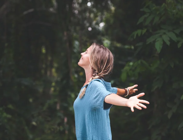 Fifty Ways to Practice Self-Care: How to Pamper Yourself for Any Occasion 5 A young woman with long hair enjoys a moment of tranquility outdoors, arms outstretched, surrounded by lush greenery.