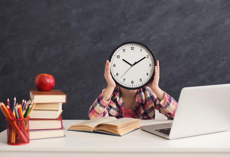 10 Habits of Successful People You Can Begin Practicing Today 6 A child holding a clock in front of their face while sitting at a desk with books, an apple, and a laptop.