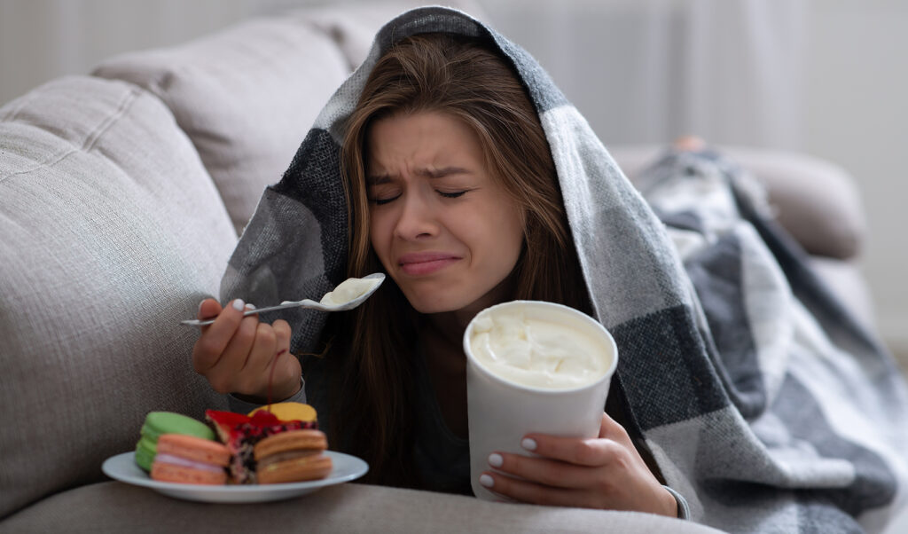15 Research-backed methods for losing weight without exercise 10 A young woman wrapped in a blanket, sitting on a couch, holding a spoonful of ice cream and a plate of colorful desserts, looking upset and emotional.