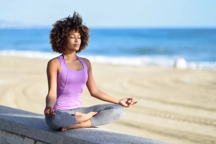 Struggling with Overthinking? 4 A young woman with curly hair practices yoga on a beach, sitting cross-legged with a serene expression against a backdrop of calm ocean waves and sandy shore.