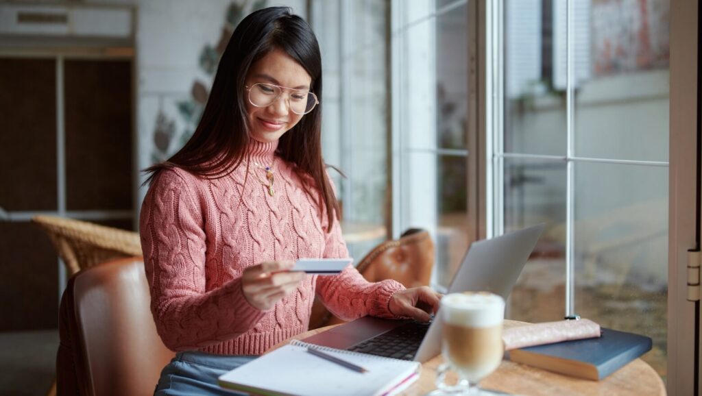 10 Habits of Successful People You Can Begin Practicing Today 7 A young woman smiles while holding a credit card in front of a laptop, with a notepad and coffee in a cozy cafe setting.