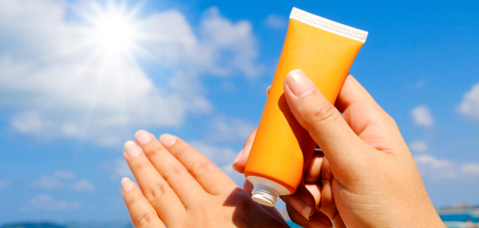 The Study of Sunscreen Science 2 A person holding a tube of sunscreen in one hand while applying it to their other hand, with a bright sun and blue sky in the background.