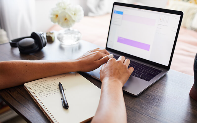 10 Habits of Successful People You Can Begin Practicing Today 4 Person typing on a laptop with a notebook and pen on a wooden desk, featuring a floral arrangement in the background.
