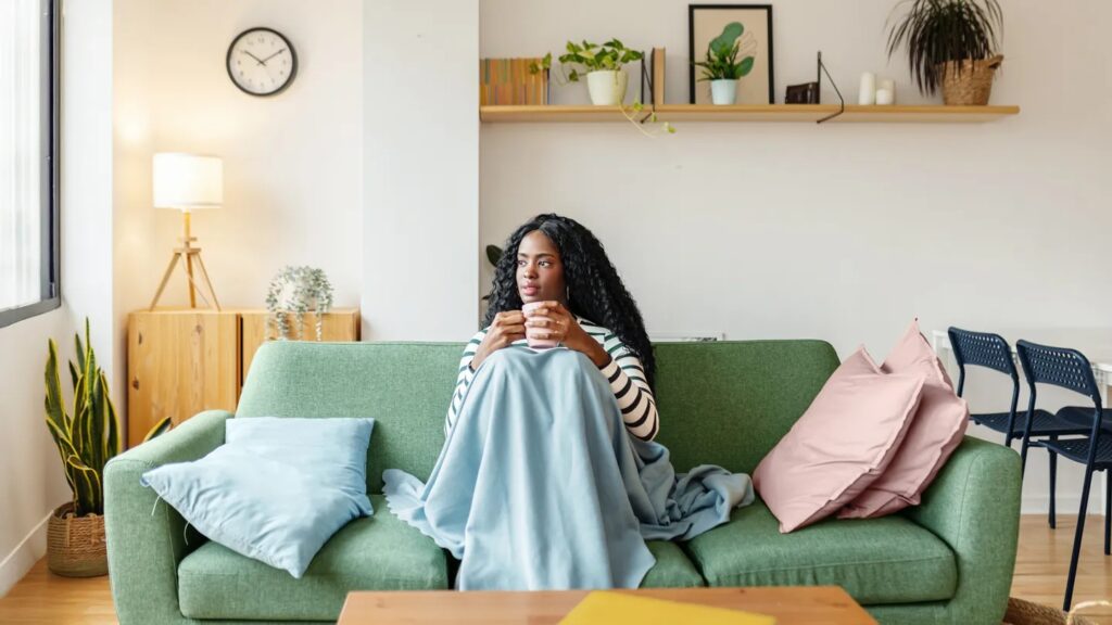 10 Habits of Successful People You Can Begin Practicing Today 5 A woman sitting on a green couch wrapped in a blanket, holding a cup, with a relaxed expression in a stylish living room.