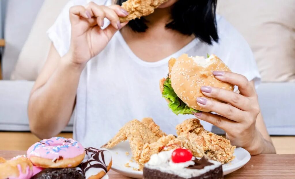 Habits to Avoid That Drain Your Energy Everyday 4 A person enjoying a variety of comfort foods, including fried chicken, a burger, and assorted desserts.
