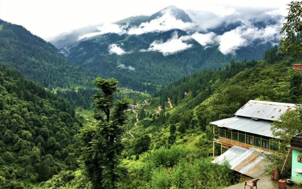 Hippie Trails in India That Promise a Journey to Freedom 3 A stunning landscape featuring rolling hills, dense forests, and misty mountains under a cloudy sky, with a rustic building in the foreground.