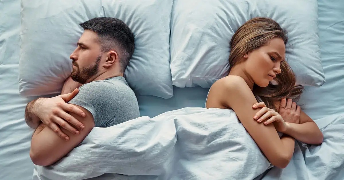 A couple lying apart in bed, facing away from each other, suggesting tension or disconnection in their relationship.
