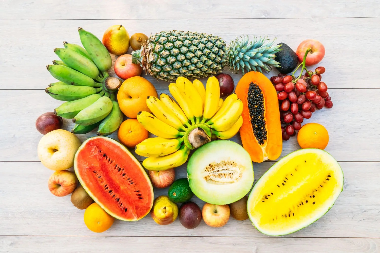 A colorful arrangement of various fresh fruits including bananas, pineapples, watermelon, papaya, grapes, and more, displayed against a light wooden background.