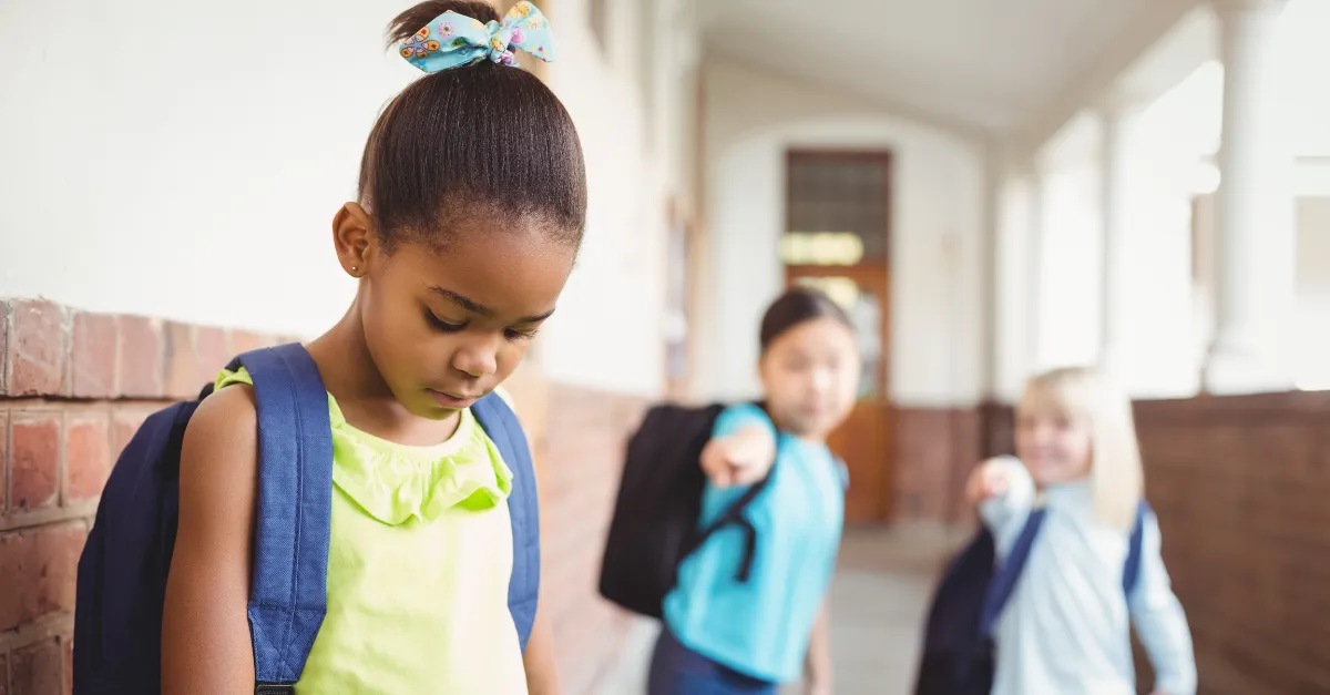 A young girl in a brightly colored shirt looks down, appearing sad or upset, while two other girls in the background point at her, highlighting a scene of bullying in a school hallway.
