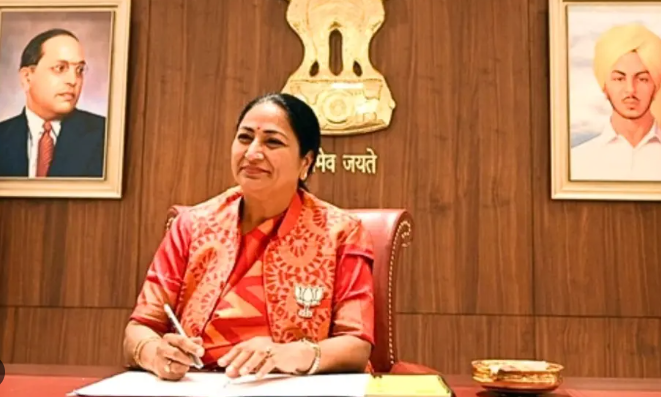 A smiling woman in a red attire sits at a desk, writing, with portraits of notable figures in the background and a state emblem above.