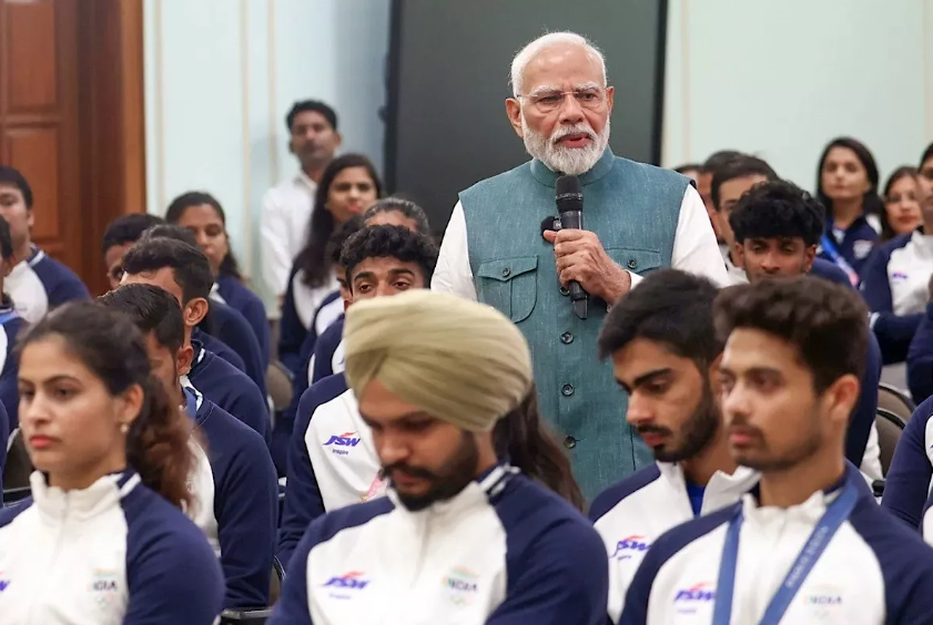 Prime Minister addressing a gathering of young athletes in a formal setting, with attentive listeners in club uniforms.