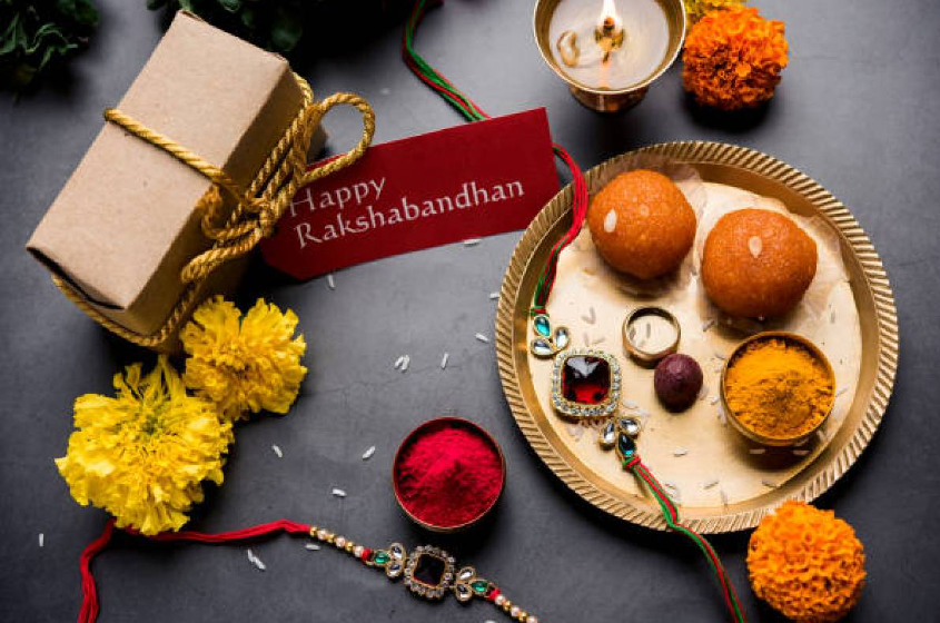 A festive arrangement featuring sweets, flowers, and a decorative plate for Rakshabandhan, alongside a gift box and greeting card.