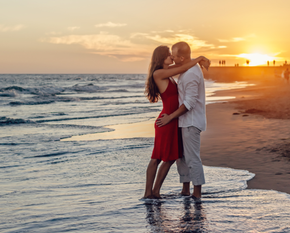 A couple shares a kiss in shallow water at sunset, with the ocean waves gently lapping around them. The woman wears a red dress, while the man is dressed in a light shirt and pants, creating a romantic atmosphere.