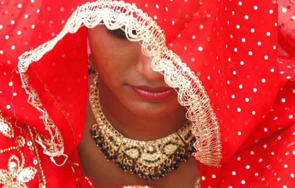 Close-up of a woman adorned in a red bridal outfit with intricate embroidery and a decorative veil, showcasing her jewelry and expression.