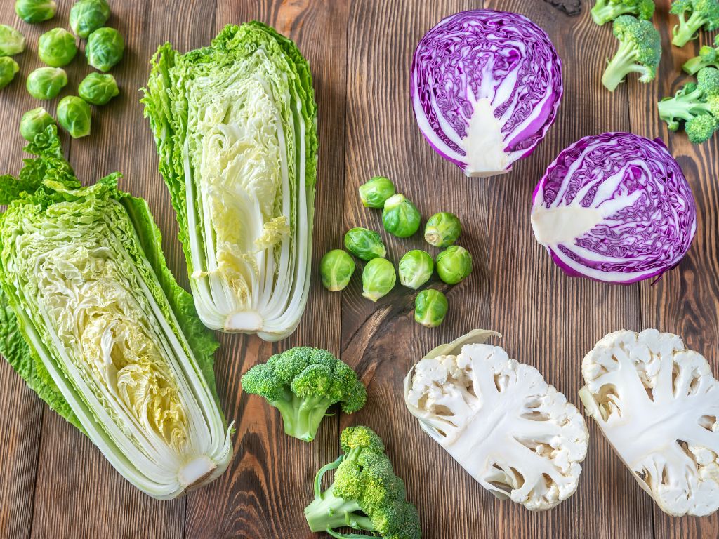 A colorful assortment of fresh cruciferous vegetables including napa cabbage, purple cabbage, broccoli, Brussels sprouts, and cauliflower displayed on a rustic wooden surface.
