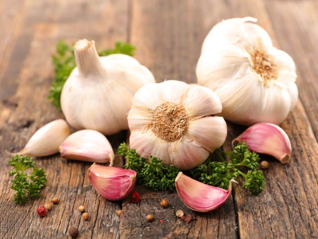 Fresh garlic bulbs and cloves arranged on a rustic wooden table with scattered herbs and spices.