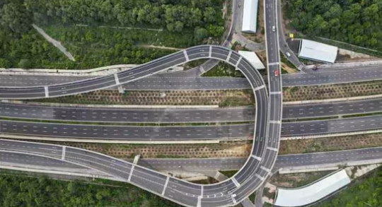 Aerial view of a complex highway interchange surrounded by lush green trees, showcasing multiple lanes and curves.