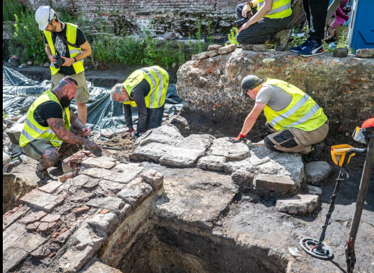 Team members excavating ancient structures at an archaeological site under sunlight, wearing safety vests and utilizing tools.