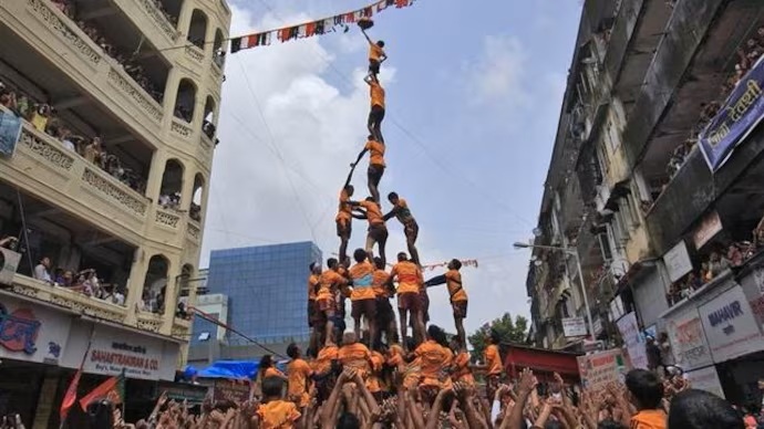 Tower of Strength: A Thrilling Human Pyramid Formation at a Festival:::A group of performers in orange shirts forms a towering human pyramid, with the top person balancing precariously while spectators cheer from below in a vibrant street festival setting.