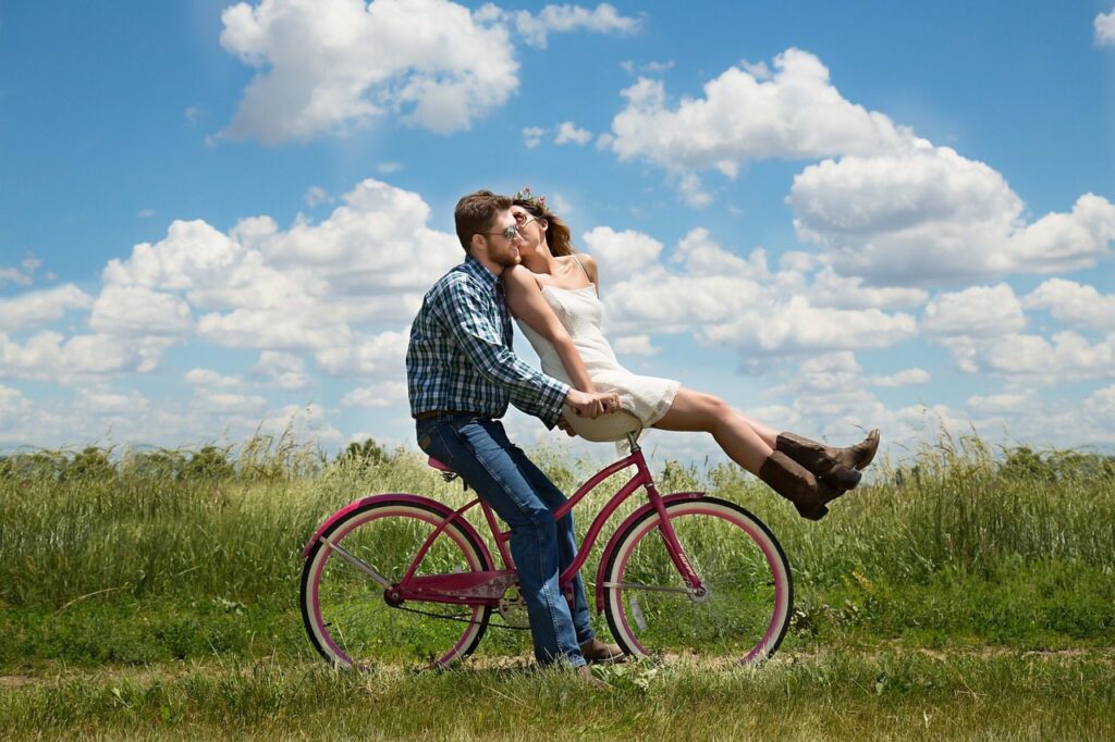 A couple sharing a kiss while riding a pink bicycle in a lush, green field under a blue sky with fluffy clouds.