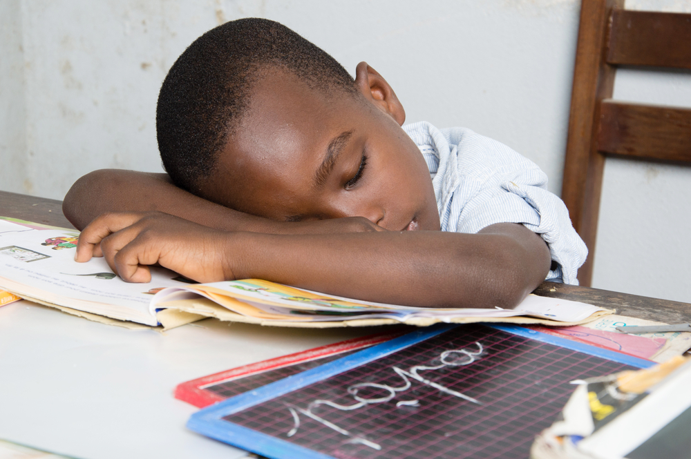 A young boy resting his head on books at a study table, surrounded by school materials and a chalkboard.