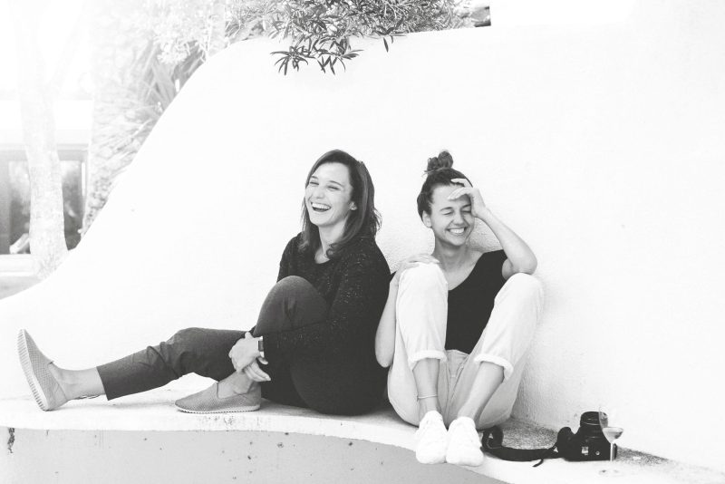 Two women sitting on a white wall, laughing joyfully while enjoying each other's company, captured in a candid black and white photo.
