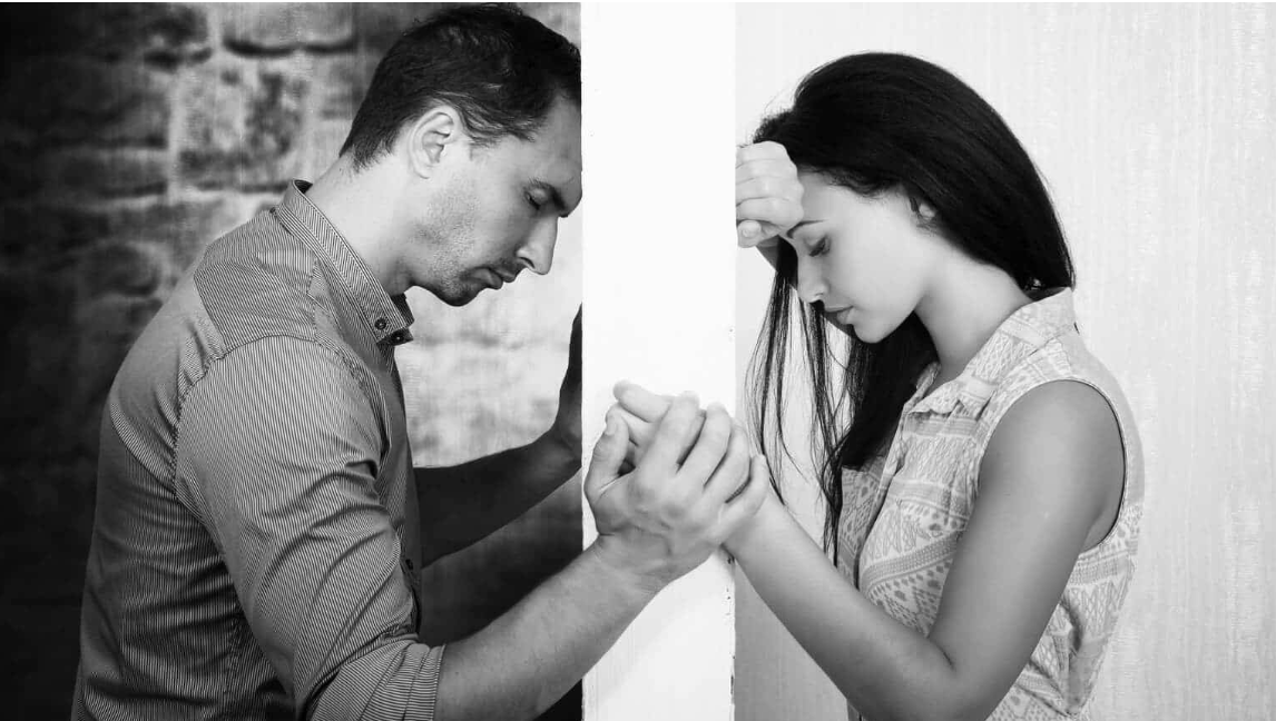 A man and woman face away from each other, expressing deep emotions as they hold hands through a wall, highlighting a moment of tension and heartache.