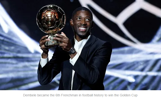 Dembele holds the Golden Cup trophy on stage, smiling widely while dressed in formal attire.